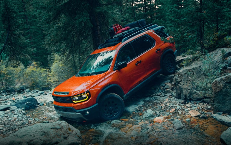2026 Honda Passport TrailSport Elite in Sunset Orange front driving over rocky terrain in a forest (Mid-Size SUV).