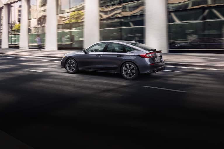 2027 Honda Civic Hatchback Sport Touring Hybrid in Meteorite Grey Metallic profile driving on a city street (Compact Sedan).